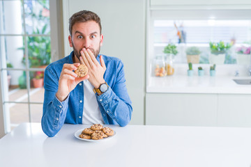 Handsome man eating chocolate chips cookies cover mouth with hand shocked with shame for mistake, expression of fear, scared in silence, secret concept