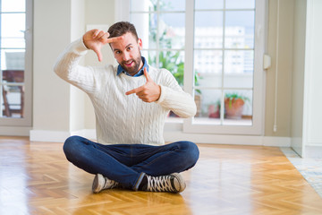 Handsome man wearing casual sweater sitting on the floor at home smiling making frame with hands and fingers with happy face. Creativity and photography concept.