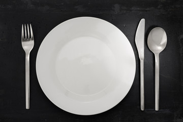 Top view table setting of an empty white plate, stainless steel silverware, isolated on black stone background with copy space.