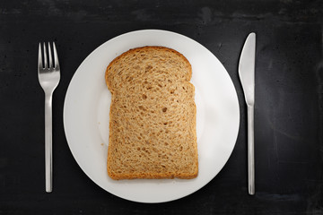 Top view of a slice of brown bread, white plate, stainless steel silverware on black stone background.