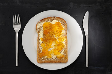 Apricot jam, slice of brown bread on white plate, stainless steel silverware and black stone background.
