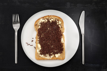 Slice of brown bread with chocolate sprinkles -or in Dutch hagelslag- on white plate, stainless steel silverware and black stone background.