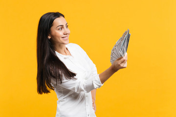 Portrait of smiling young woman in white casual shirt holding fan of cash money in dollar banknotes isolated on yellow orange wall background in studio. People lifestyle concept. Mock up copy space.