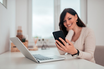 Beautiful happy woman using smart phone and laptop at home office, focus on the foreground.