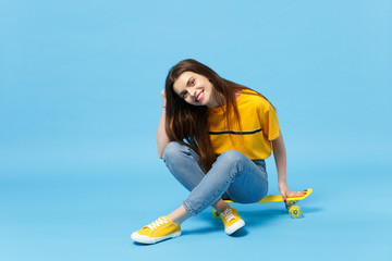 Portrait of pretty cute young woman in vivid casual clothes looking camera and sitting with yellow skateboard isolated on blue wall background in studio. People lifestyle concept. Mock up copy space.