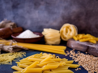 Italian pasta, spaghetti, fettuccine, penne, rolling pin, flour on a textured background. Still life in a rustic style.