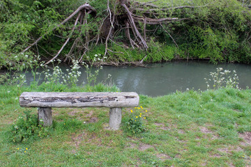 Fototapeta na wymiar An empty old wooden bench in front of a clean stream. Location is the river Windrush in the Cotswolds countryside, Witney, Oxfordshire, UK.