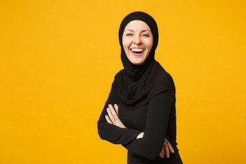 Smiling young arabian muslim woman in hijab black clothes hold hands folded, looking camera isolated on yellow wall background, studio portrait. People religious lifestyle concept. Mock up copy space.