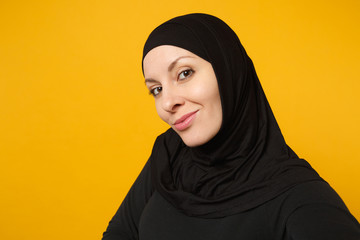 Close up young arabian muslim woman in hijab black clothes doing selfie shot on mobile phone isolated on yellow wall background studio portrait. People religious lifestyle concept. Mock up copy space.