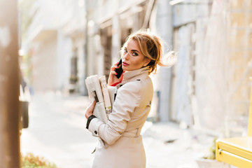 Busy attractive lady with elegant hairstyle looking over shoulder during phone conversation. Serious young woman in trendy coat hurrying to office with fresh newspapers.