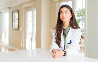 Young woman wearing medical coat at the clinic as therapist or doctor smiling looking side and...