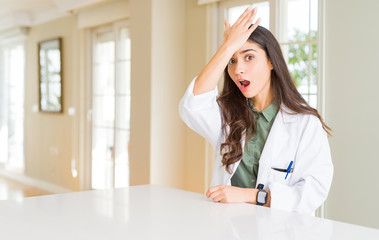 Young woman wearing medical coat at the clinic as therapist or doctor surprised with hand on head...