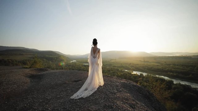 Young beautiful woman in wedding dress walks in mountains at sunset. Back view