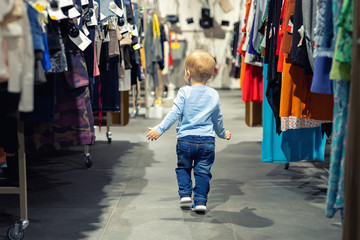 Cute caucasian blond toddler boy walking alone at clothes retail store between rack with hangers....