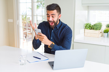 Handsome hispanic business man using smartphone and laptop at the office very happy and excited, winner expression celebrating victory screaming with big smile and raised hands