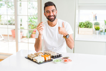 Handsome hispanic man eating asian sushi using chopsticks happy with big smile doing ok sign, thumb up with fingers, excellent sign