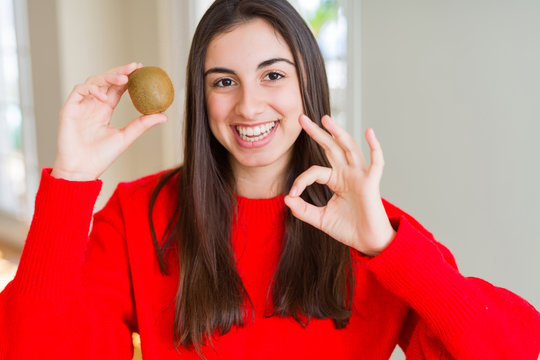 Beautiful young woman eating fresh green kiwi doing ok sign with fingers, excellent symbol
