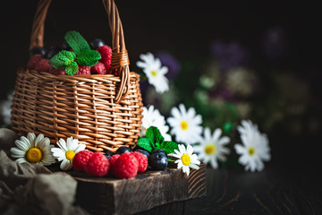 Fototapeta na wymiar Raspberries and blueberries in a basket with chamomile and leaves on a dark background. Summer and healthy food concept. Background with copy space. Selective focus.
