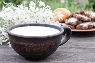 Next to the cookies and white flowers is a cup of milk on a wooden table.