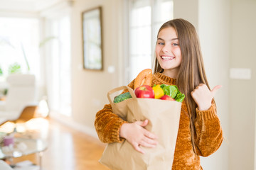 Beautiful young girl holding paper bag of fresh groceries pointing and showing with thumb up to the side with happy face smiling