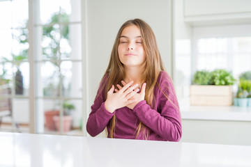 Beautiful young girl kid on white table smiling with hands on chest with closed eyes and grateful gesture on face. Health concept.