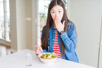 Beautiful Asian woman eating healthy pasta salad cover mouth with hand shocked with shame for mistake, expression of fear, scared in silence, secret concept