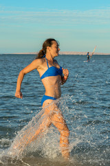A sexy young brunette woman or girl wearing a bikini running through the surf on a deserted tropical beach with a blue sky. Young woman running by the sea. vertical photo