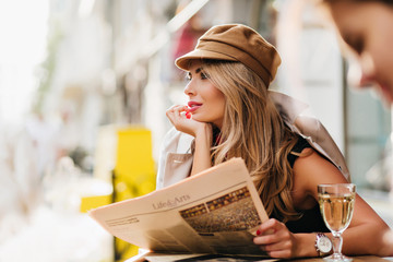Dreamy woman in brown cap thinking about something, propping face with hand and holding newspaper....