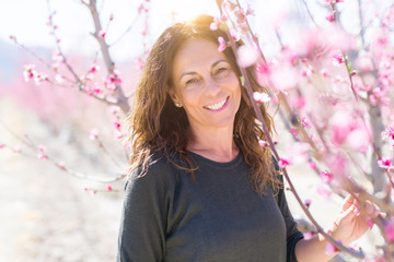 Beautiful middle age woman in the middle of pink peach flowers and trees smiling cheerful, colorful...