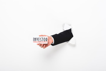 Cropped view of man holding card with investor word through hole in paper wall on white