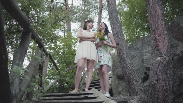 Two young women in short dreses with flowers standing on the stairs and looking at amzing forst, trees, plants. Pretty girls walk outside the city. Cute girlfriends spending weekend together outdoors.
