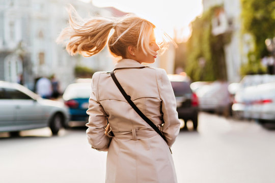 Slim Woman In Stylish Coat Walking Down The Street Alone And Looking Around. Outdoor Portrait From Back Of Wonderful Blonde Lady Exploring City In Sunny Day.