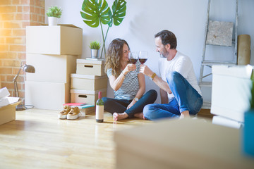 Middle age senior romantic couple in love sitting on the apartment floor with boxes around, celebrating drinking a glass of wine smiling happy for moving to a new home