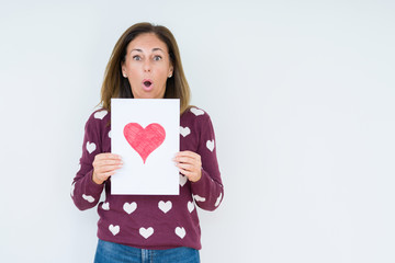 Middle age woman holding card gift with red heart over isolated background scared in shock with a surprise face, afraid and excited with fear expression