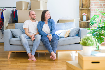 Young couple sitting on the sofa arround cardboard boxes moving to a new house looking away to side with smile on face, natural expression. Laughing confident.