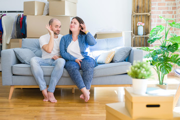 Young couple sitting on the sofa arround cardboard boxes moving to a new house smiling with hand over ear listening an hearing to rumor or gossip. Deafness concept.