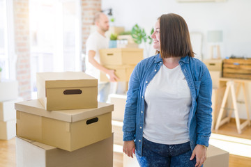 Fototapeta na wymiar Young couple arround cardboard boxes moving to a new house, plus size woman standing at home looking to side, relax profile pose with natural face with confident smile.