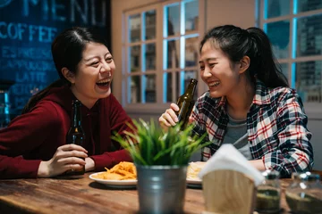 Foto op Plexiglas romantic asian lesbian couple drinking beer in bottle at vintage bar in modern city. young girls laughing chatting in ladies night in pub enjoy alcohol get drunk sitting at wood table with chips. © PR Image Factory