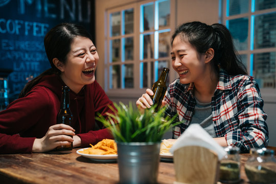 Romantic Asian Lesbian Couple Drinking Beer In Bottle At Vintage Bar In Modern City. Young Girls Laughing Chatting In Ladies Night In Pub Enjoy Alcohol Get Drunk Sitting At Wood Table With Chips.