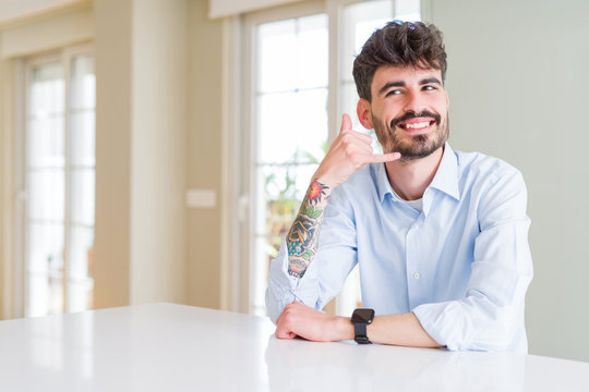 Young businesss man sitting on white table smiling doing phone gesture with hand and fingers like talking on the telephone. Communicating concepts.