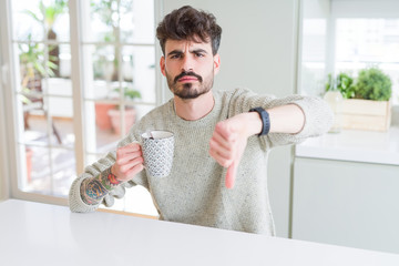 Young man drinking a cup of coffee in the morning with angry face, negative sign showing dislike...