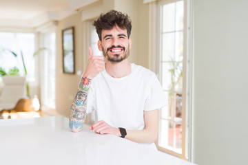 Fototapeta premium Young man wearing casual t-shirt sitting on white table doing happy thumbs up gesture with hand. Approving expression looking at the camera showing success.