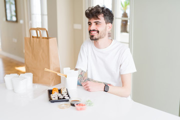 Young man eating asian sushi from home delivery looking away to side with smile on face, natural expression. Laughing confident.