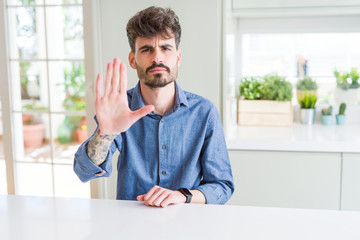 Young man wearing casual shirt sitting on white table doing stop sing with palm of the hand. Warning expression with negative and serious gesture on the face.