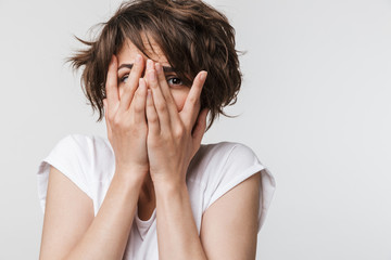 Photo of frightened woman with short brown hair in basic t-shirt keeping hands over her face and looking at camera