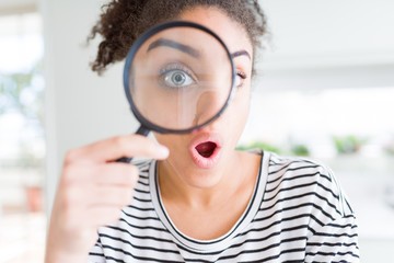 Young african american woman looking through magnifying glass scared in shock with a surprise face,...