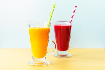 Two colorful cocktails, orange and red, in glass cups with straw. Healthy smoothies, yellow and white background.