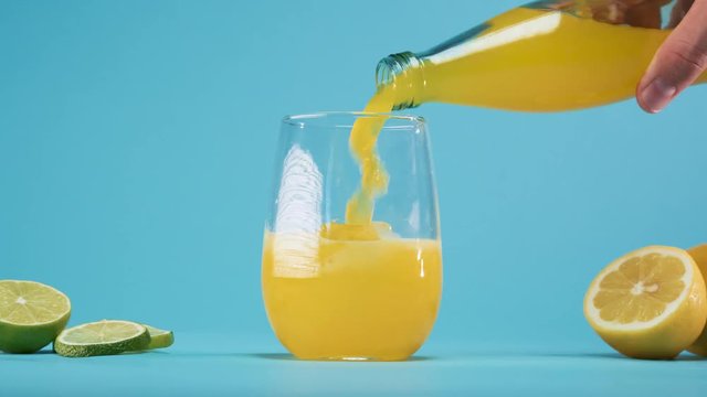 Hand holding bottle and pouring fresh and cold yellow soda lemonade drink in to the glass with ice cubes placed on blue background.