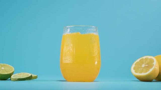 Hand places glass of fresh and cold yellow soda lemonade drink with ice cubes on blue background.