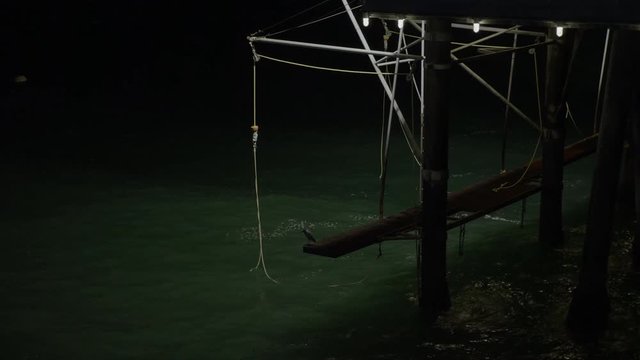 Spooky, dark, illuminated legs emerged beneath seascape waves lit with eerie glow at nighttime. Ropes hanging from structure.